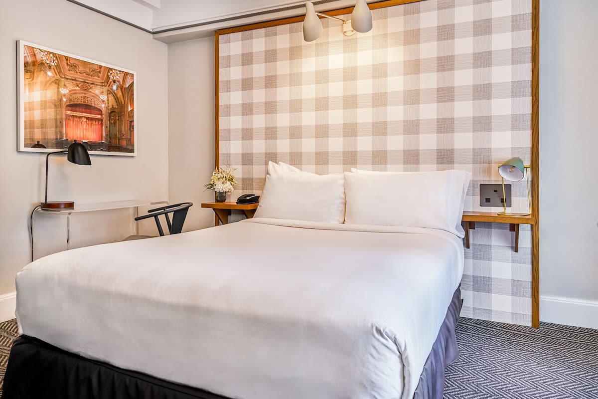 Quarto de hotel com uma cama grande, cabeceira xadrez, um quadro de um teatro, uma escrivaninha com luminária e mesa de cabeceira com luminária.