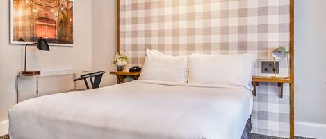 Una habitación de hotel con una cama grande, un cabecero a cuadros, un cuadro de un teatro, un escritorio con una lámpara y una mesita de noche con una lámpara.