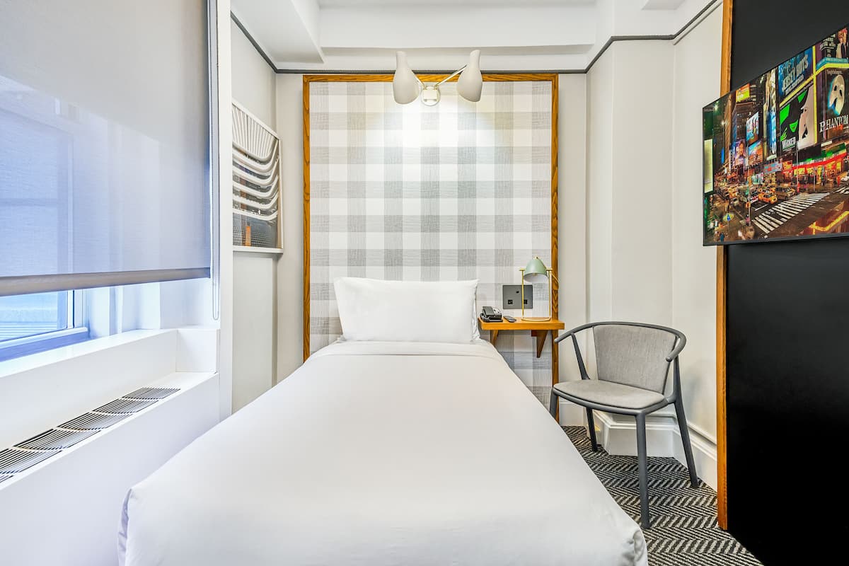 Quarto de hotel com cama, poltrona, escrivaninha com luminária, quadro e janela com persianas.