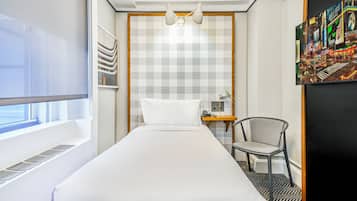 A hotel room with a bed, a chair, a desk with a lamp, a painting, and a window with blinds.