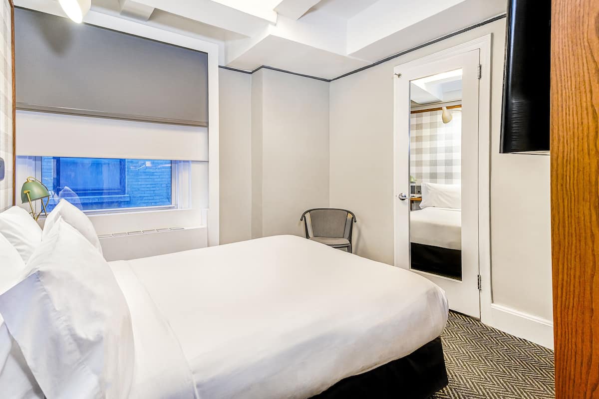 Quarto de hotel com uma cama grande, uma cadeira, uma janela com persianas, uma porta que dá para um banheiro e uma TV de tela plana na parede.