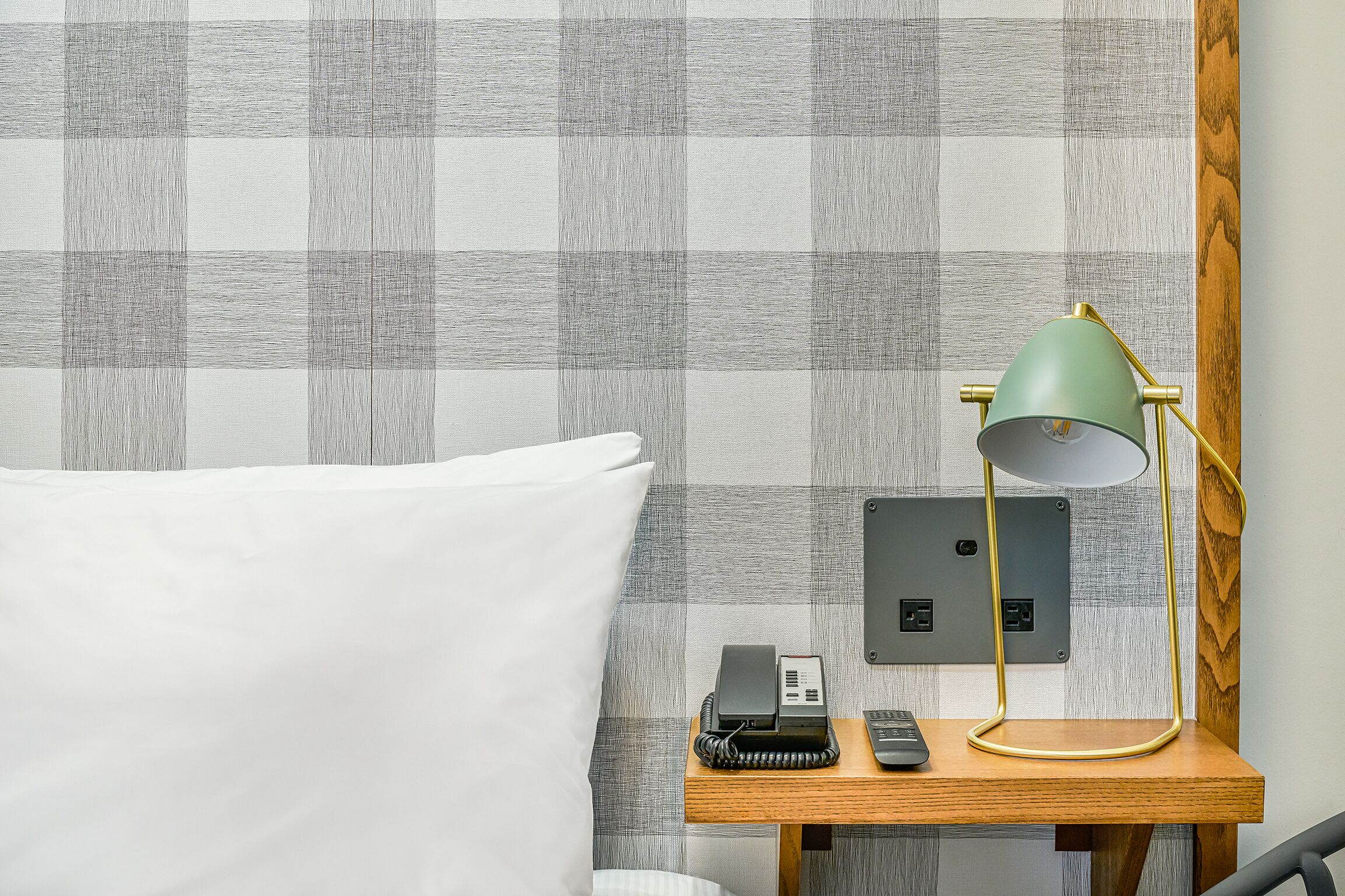 A neatly made bed with a white pillow, a wooden bedside table with a green desk lamp, a phone, and a power strip.