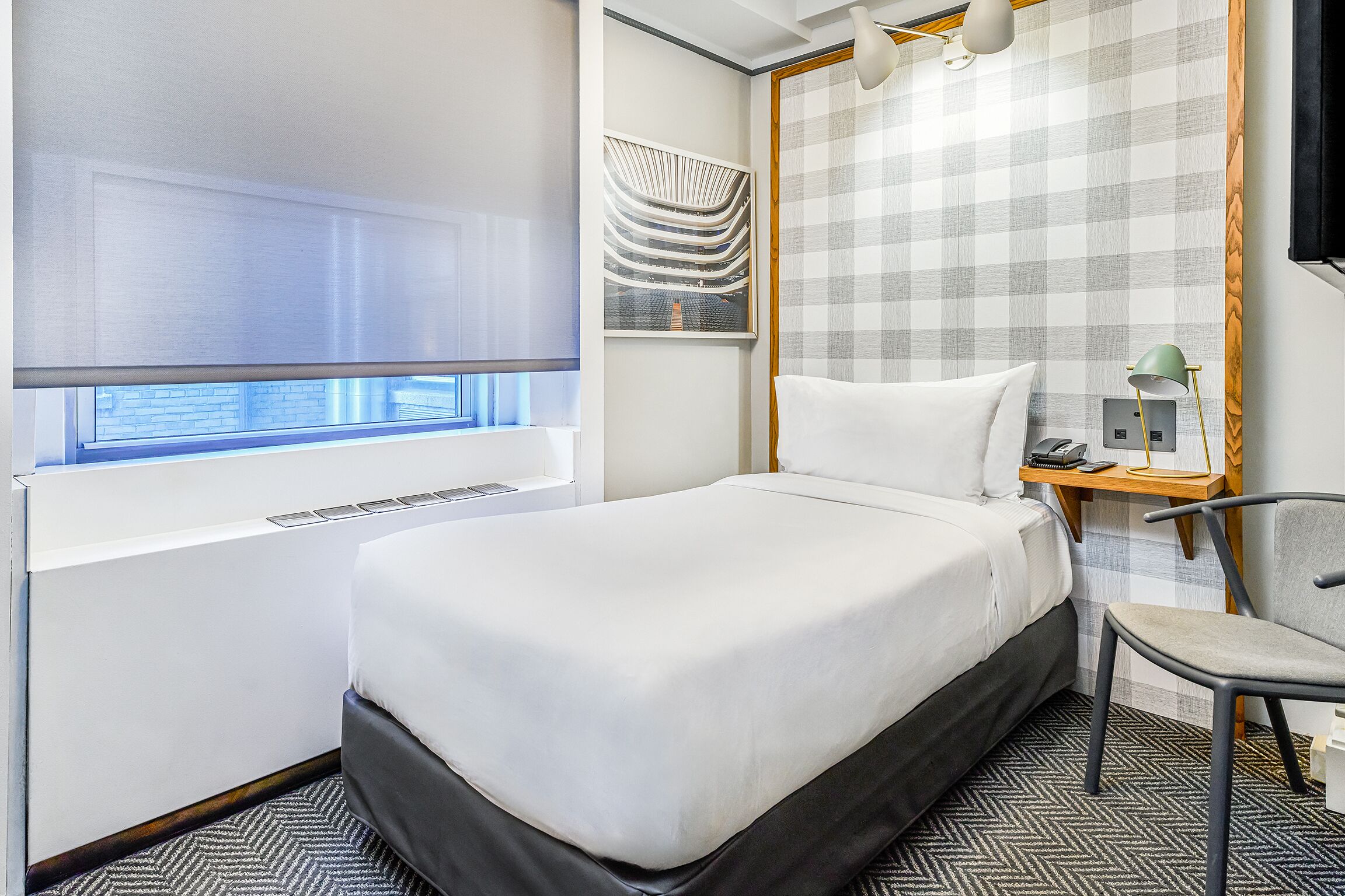 Una habitación de hotel con cama, escritorio con lámpara, silla y ventana con persianas.