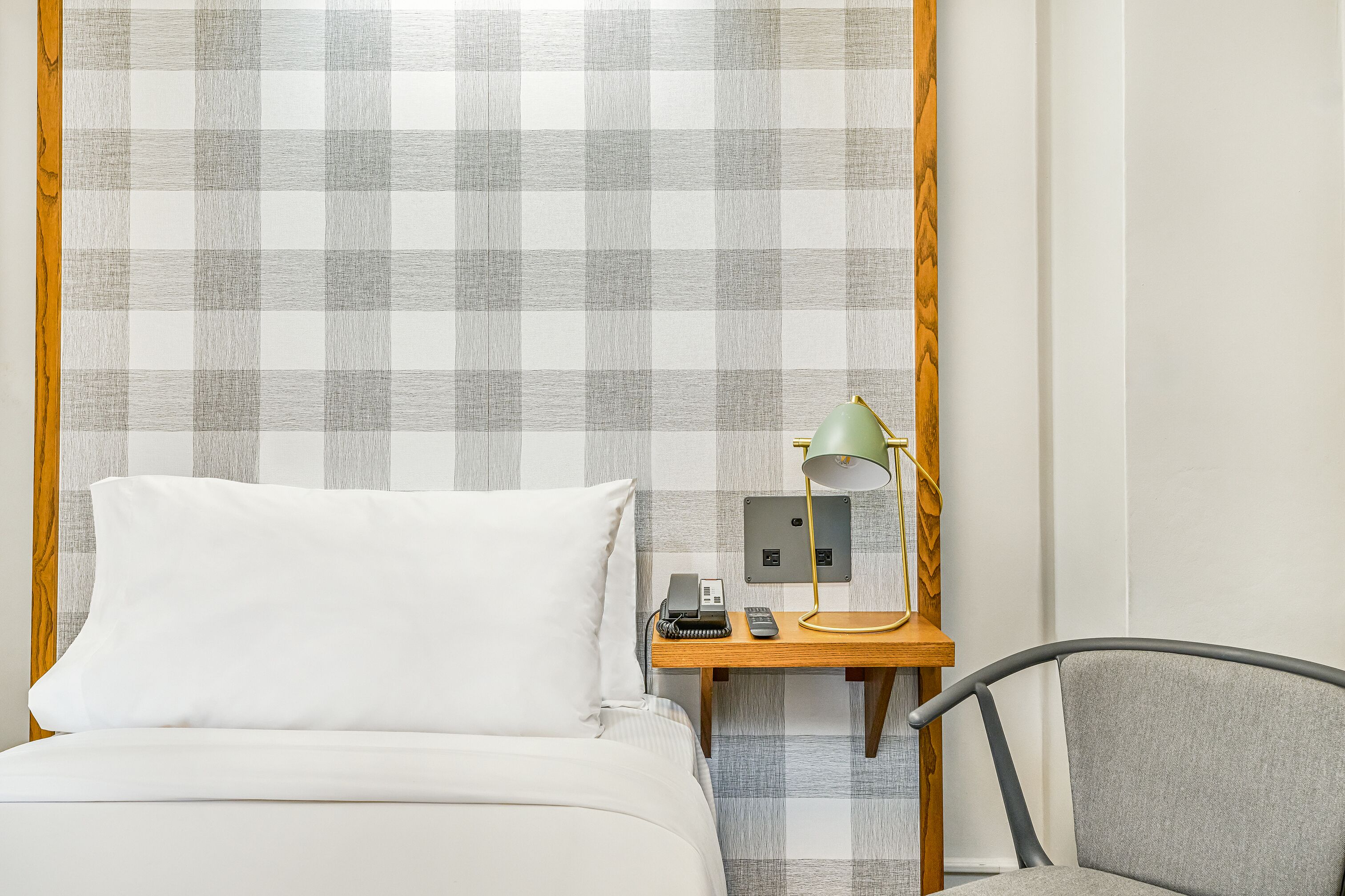Una cama bien hecha con un almohadón blanco y una mesita de noche de madera al lado. En la mesita hay una lámpara, un teléfono y una pequeña mesa.