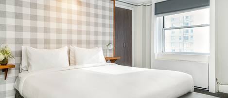 Une chambre d’hôtel avec un grand lit, une fenêtre donnant sur des immeubles, un mur à motifs à carreaux et une table de chevet sur laquelle est posé un vase de fleurs.