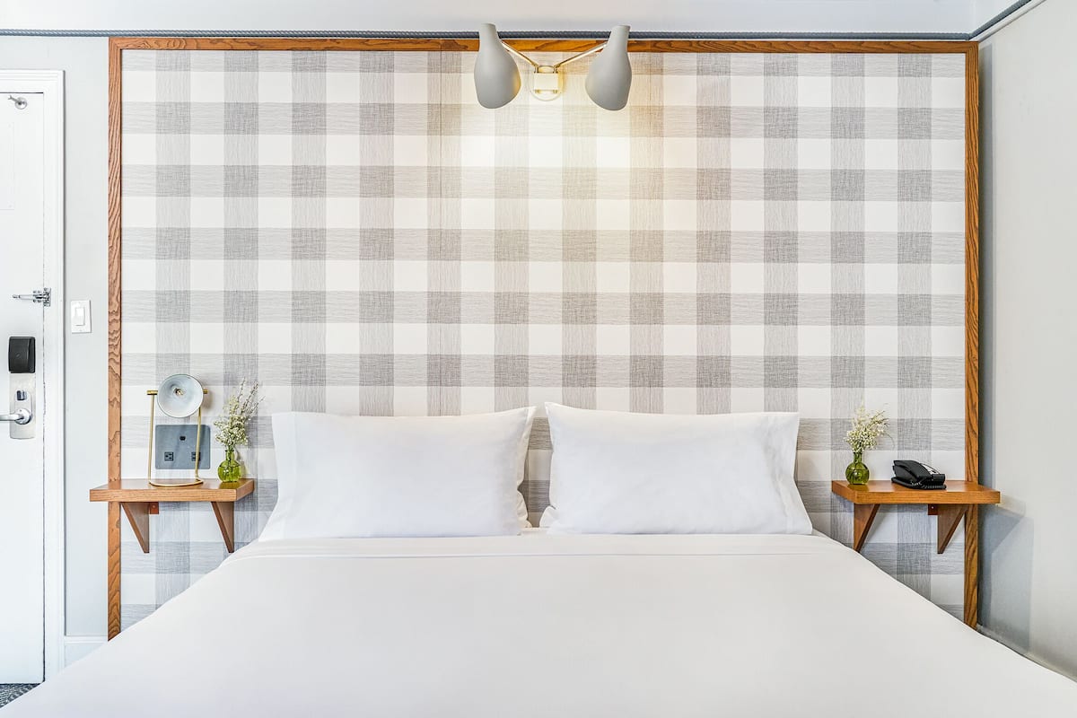 Uma cama bem arrumada com duas almofadas, uma mesa de cabeceira com abajur, telefone e um pequeno vaso com flores.