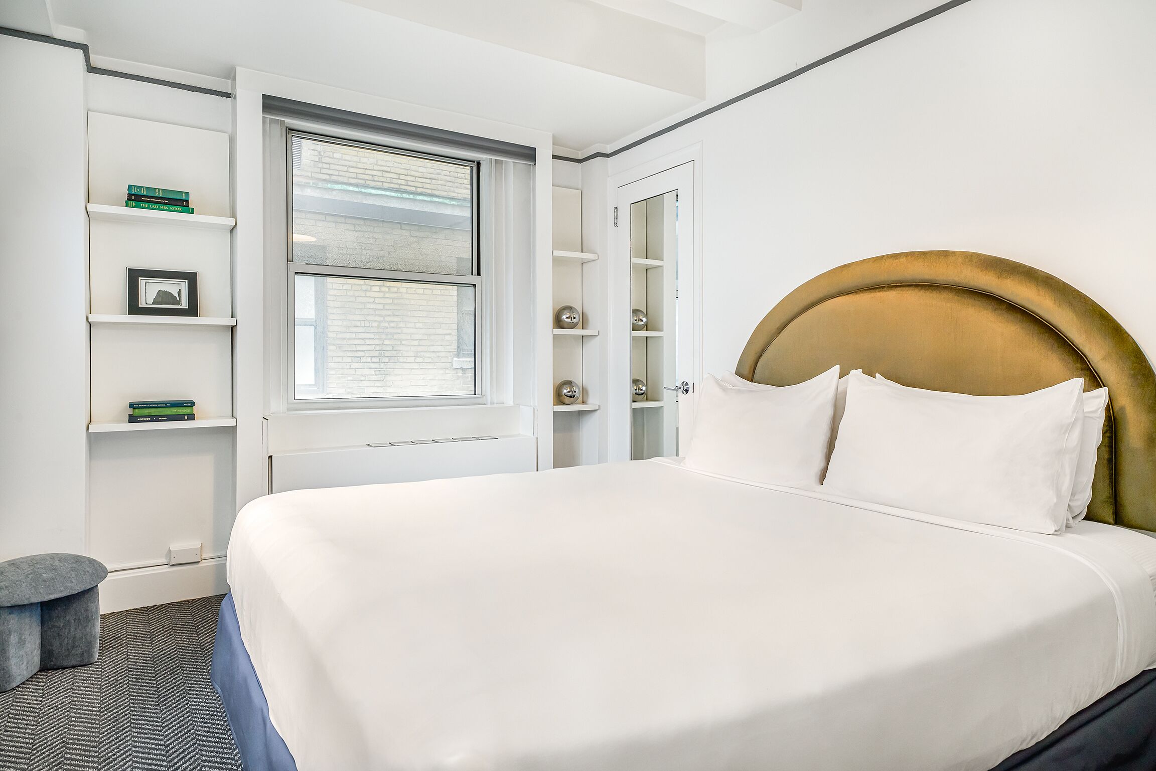 A neatly arranged hotel room with a large bed, built-in shelving with books and decorative items, a window with blinds, and a small stool.