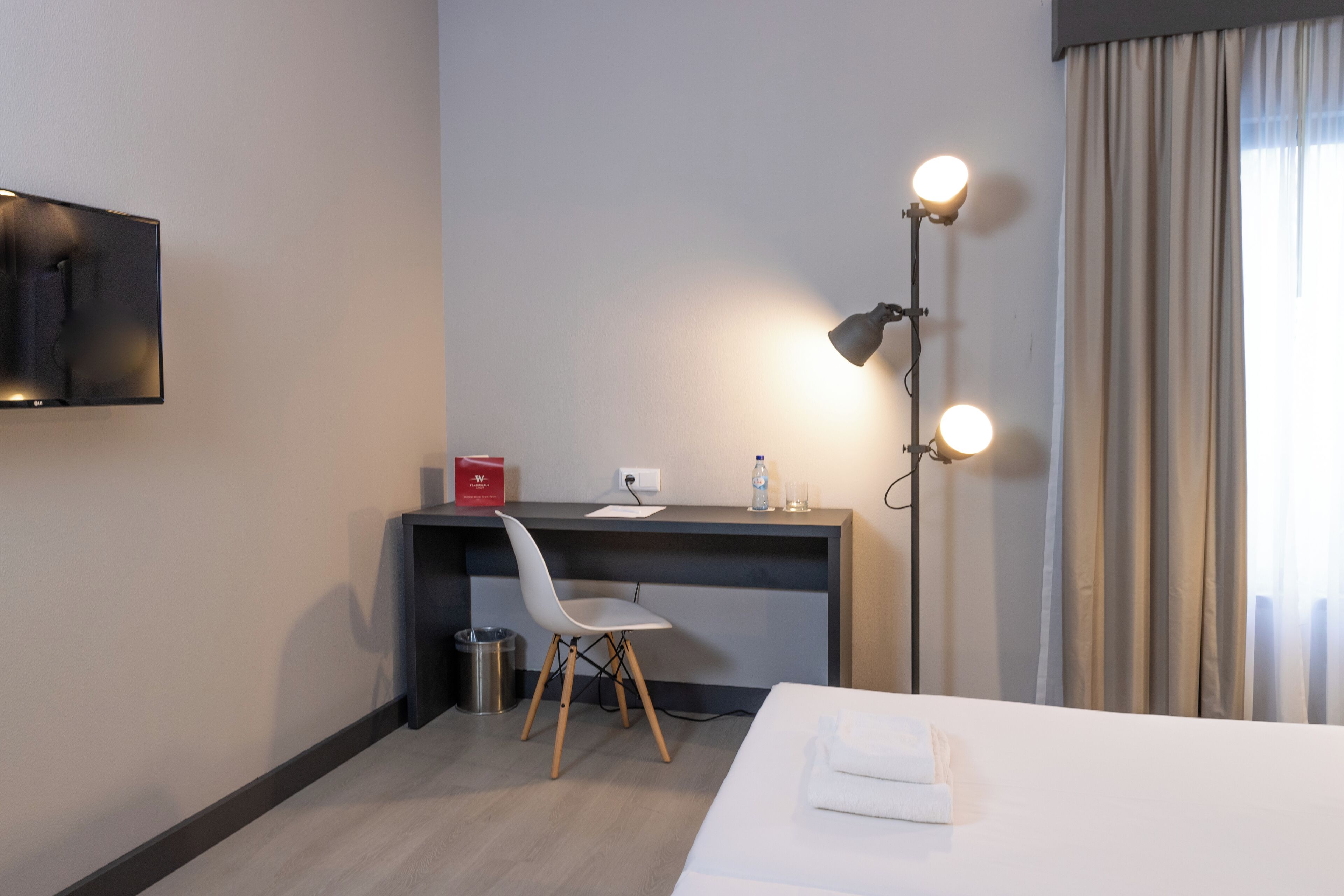 A hotel room with a bed, a desk with a chair, a desk lamp, a TV, and a window with curtains.
