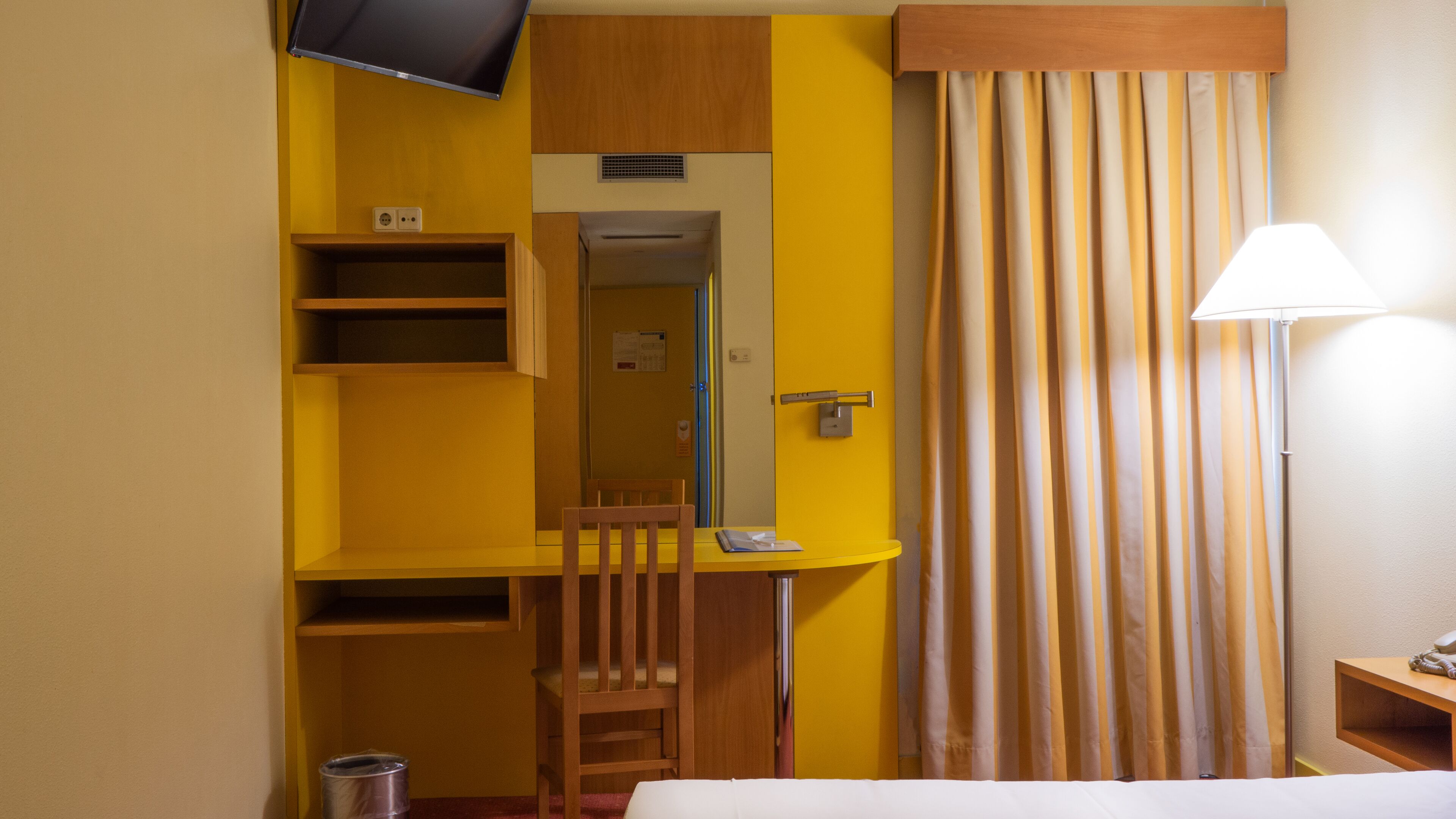 A compact hotel room with a bed, a desk, a chair, a television, a lamp, and a curtain.