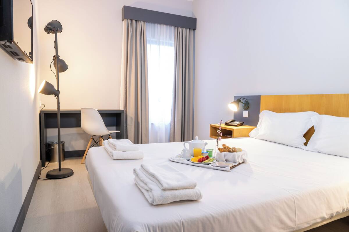 A hotel room with a neatly made bed, a bedside table with a lamp, a tray with breakfast items, and a window with curtains.