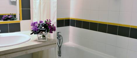 Un baño con un lavamanos blanco, un florero de vidrio con flores moradas, una ducha y un perchero para toallas.