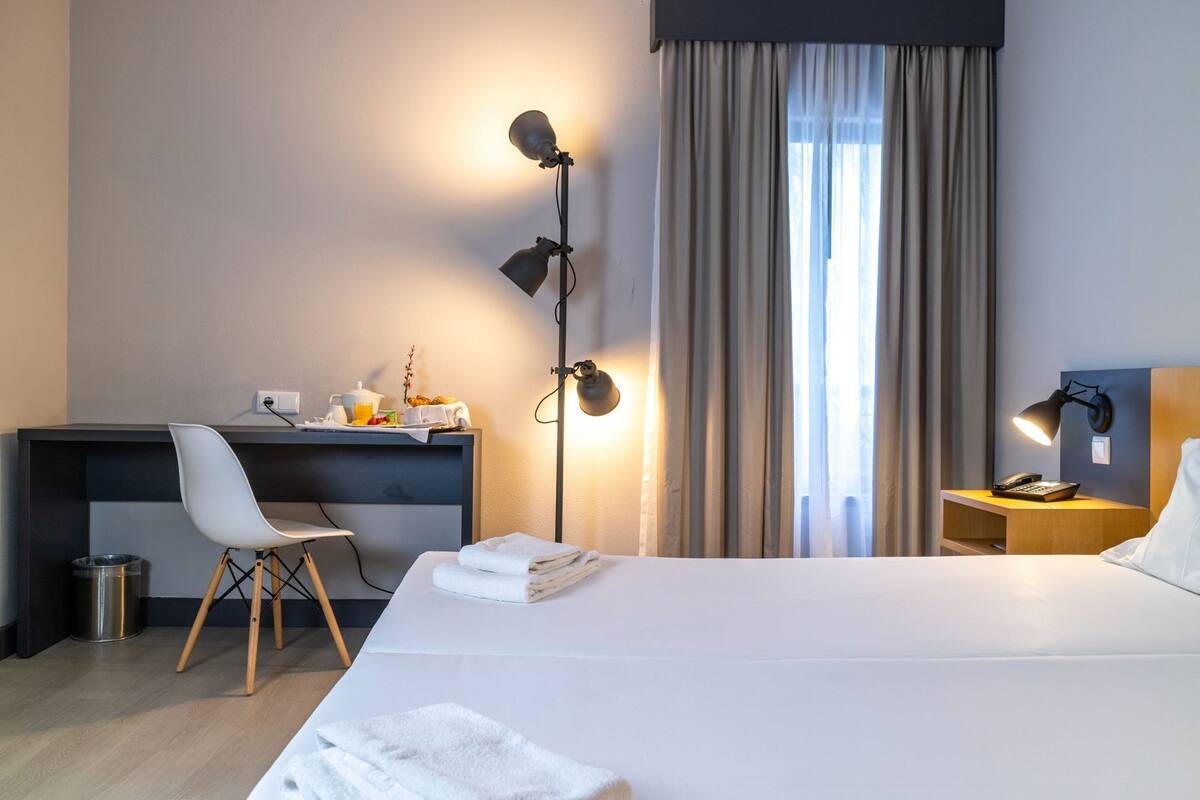 A hotel room with a bed, desk, chair, bedside table, lamp, and curtains.