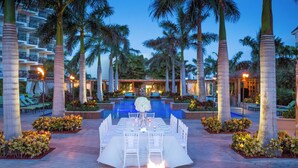 A poolside dining area with a long table set for a meal, surrounded by palm trees and a building with balconies. - Aruba Marriott Resort & Stellaris Casino (Noord)