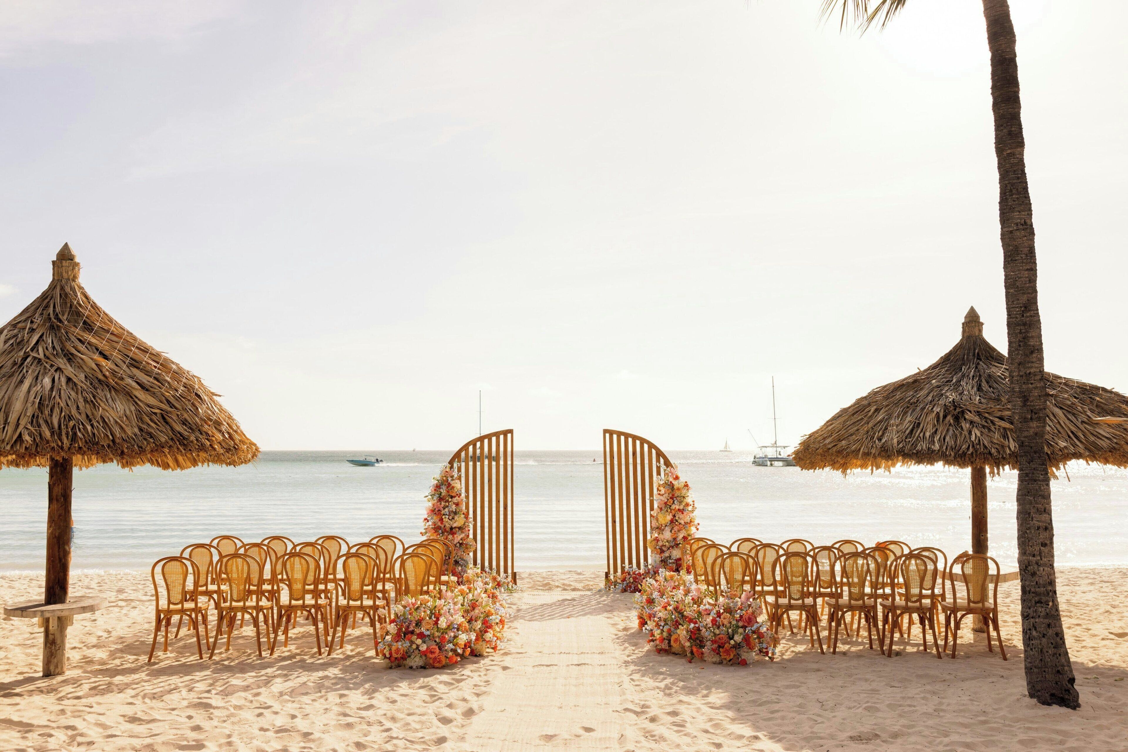 Een strandbruiloft decoratie, met een pad naar een houten boog, omringd door rieten parasols en rieten stoelen.