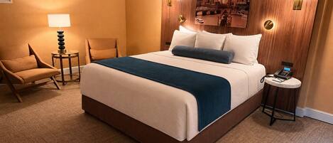 A hotel room with a large bed, a chair, a small table with a lamp, and a wall-mounted painting.