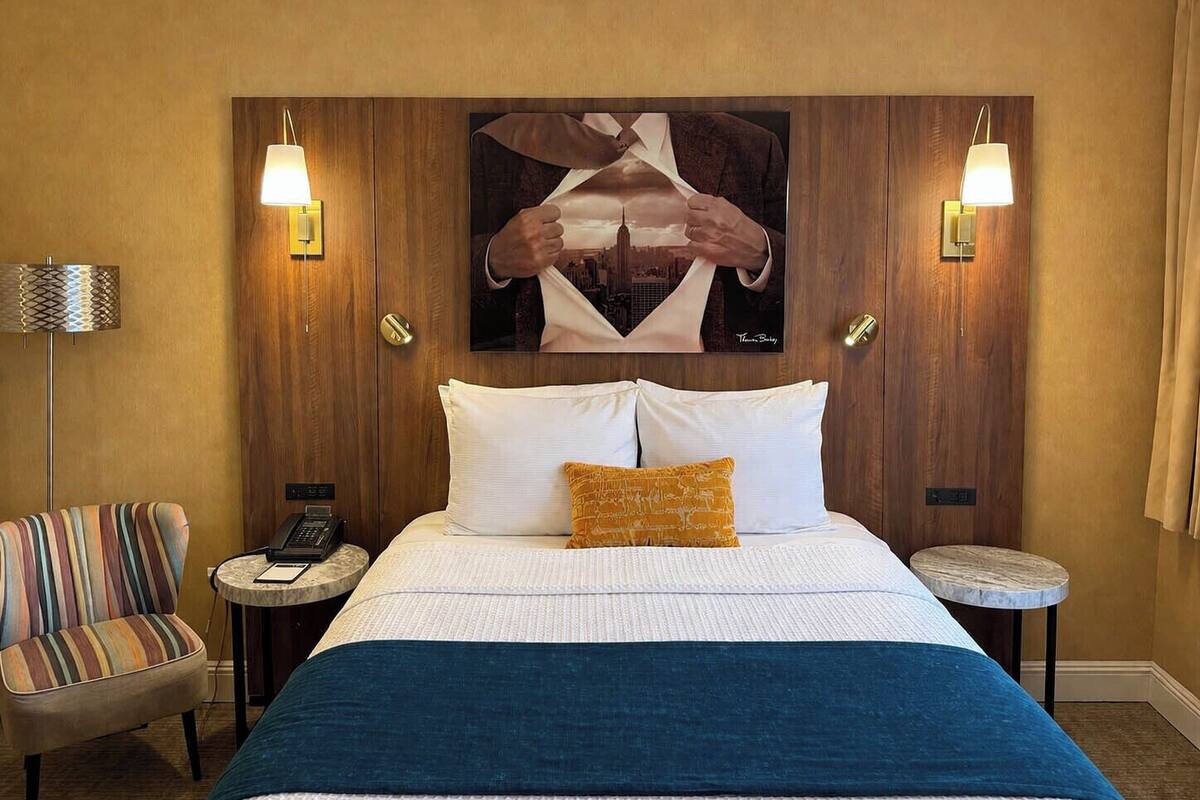 A hotel room with a large bed, two bedside tables, a striped armchair, and a cityscape artwork on the wall.
