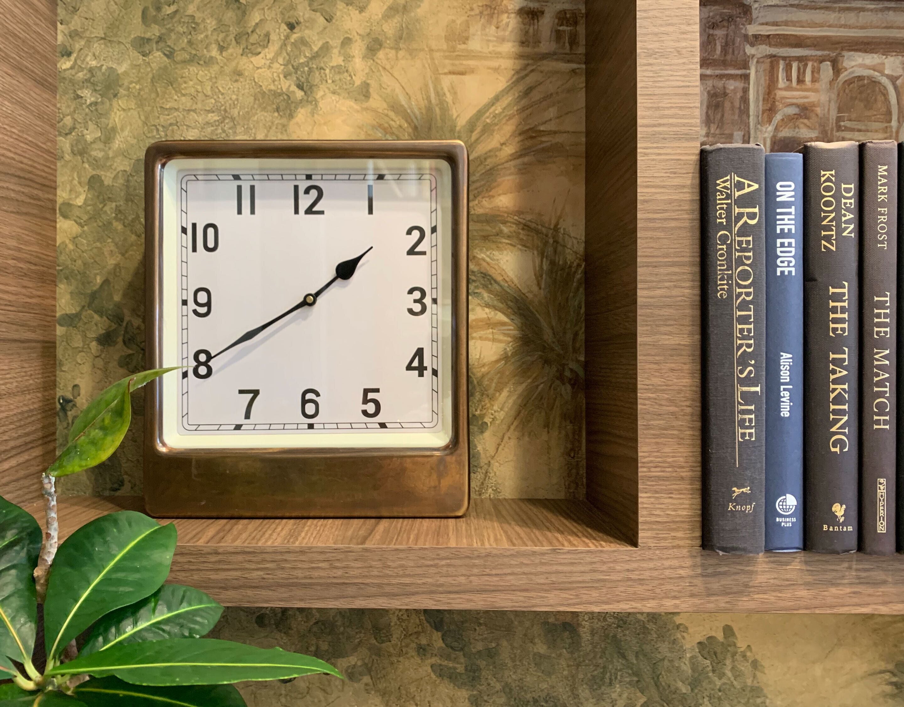 Una estantería de madera con un reloj cuadrado que marca la hora, una planta y una fila de libros.