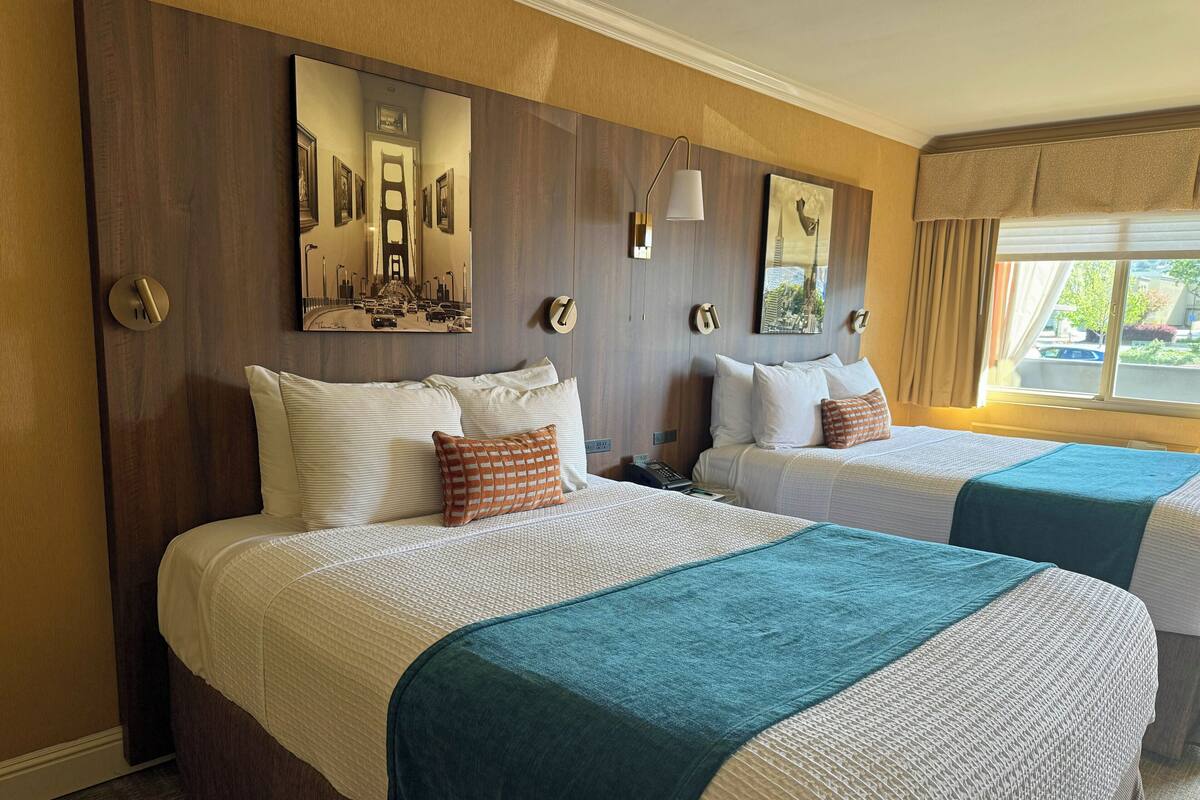 A hotel room with two beds, a large mirror, artwork on the walls, and a window with curtains.