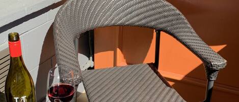 A wicker chair with a curved backrest, a bottle of wine, a glass of red wine, and a bowl of nuts on a table.
