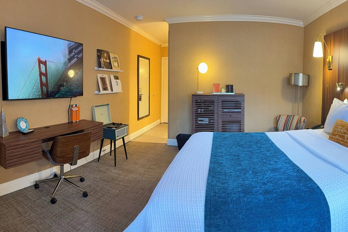 A hotel room with a bed, a desk, a chair, a TV, and a large painting on the wall.
