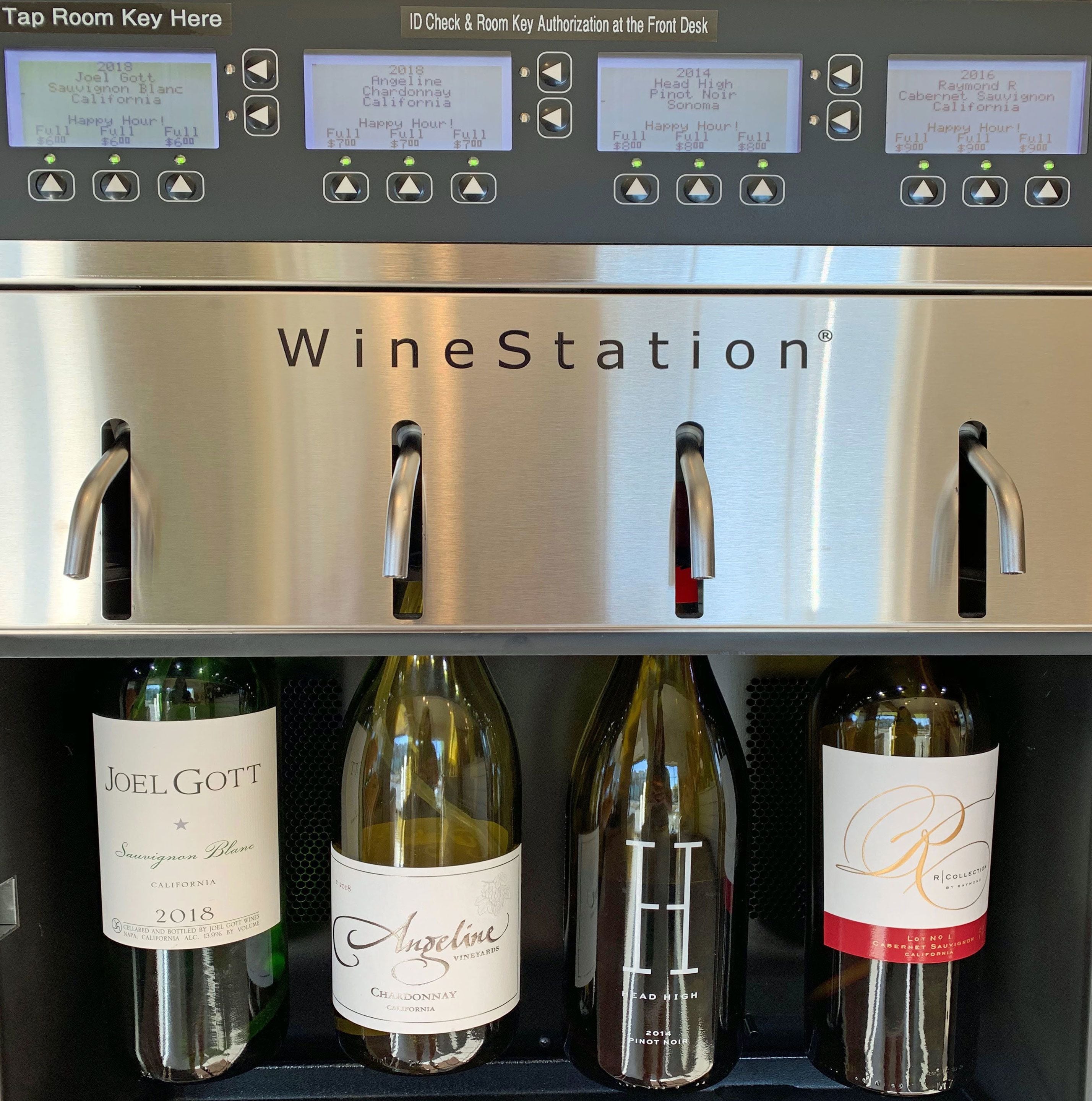Máquina dispensadora de vino con cuatro etiquetas de botellas visibles: Joel Gott Sauvignon Blanc, Angeline Chardonnay, Head High Pinot Noir y Raymond R Cabernet Sauvignon.