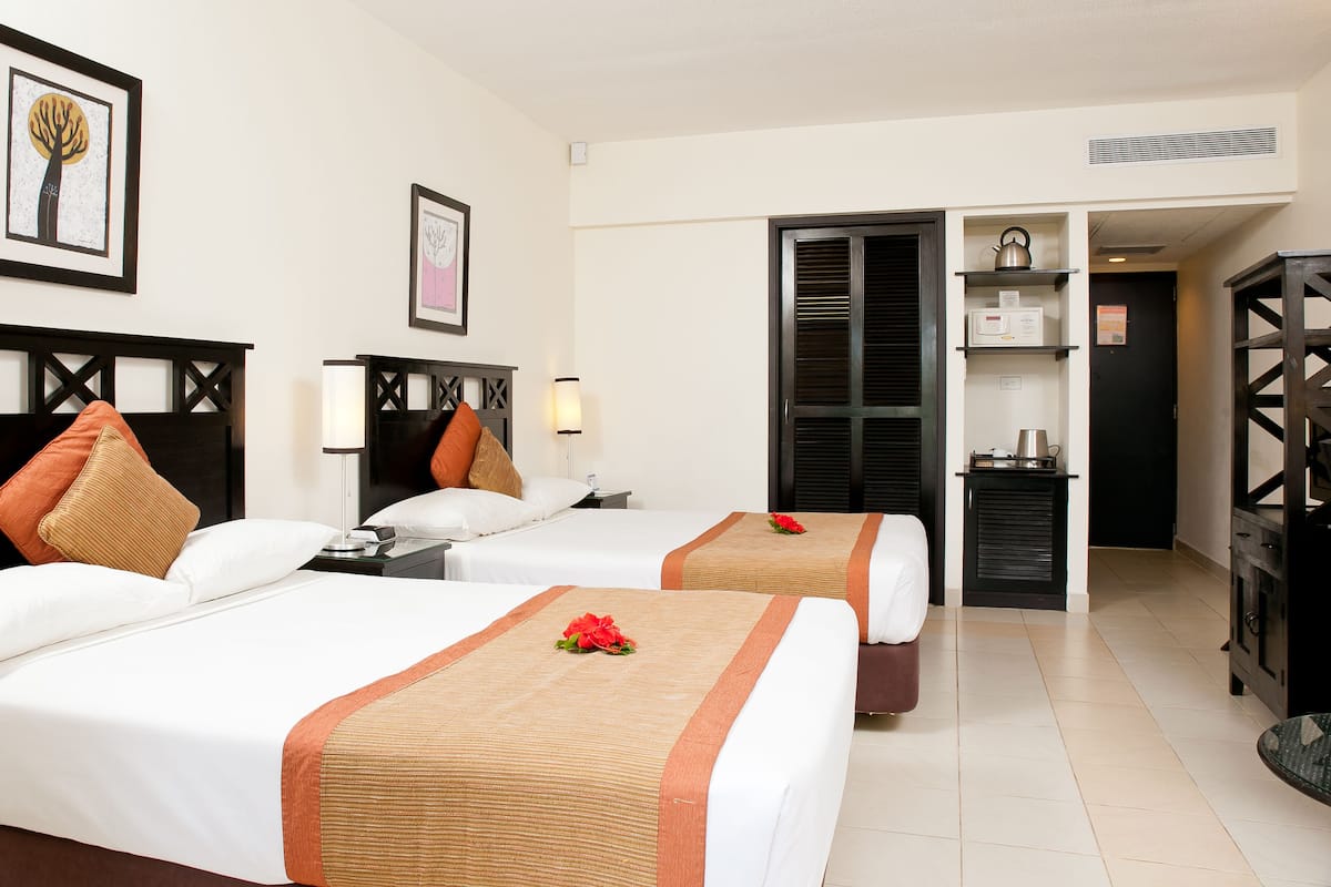 Habitación de hotel con dos camas, una mesita de noche con lámpara, un cuadro en la pared y una kitchenette.