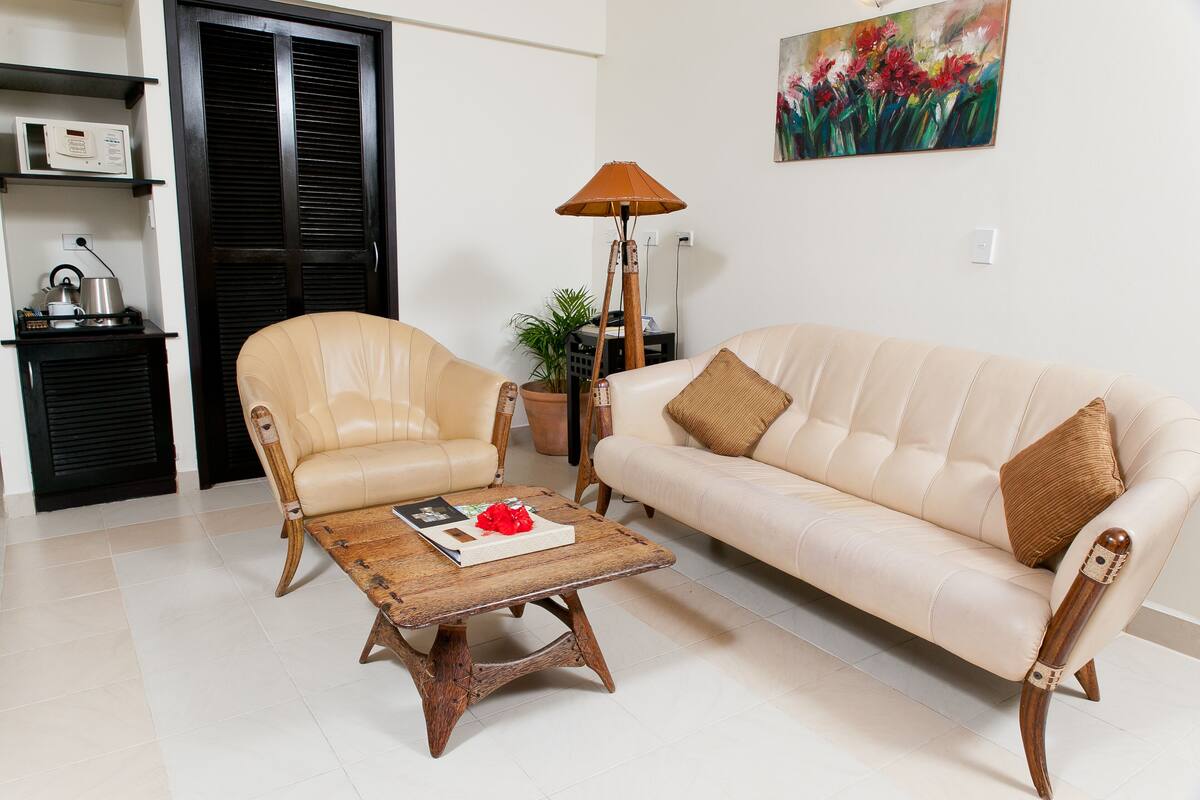 Sala de estar con un sofá color beige, un sillón a juego y una mesa de centro de madera. Hay una lámpara de pie, una planta en maceta y un cuadro en la pared.