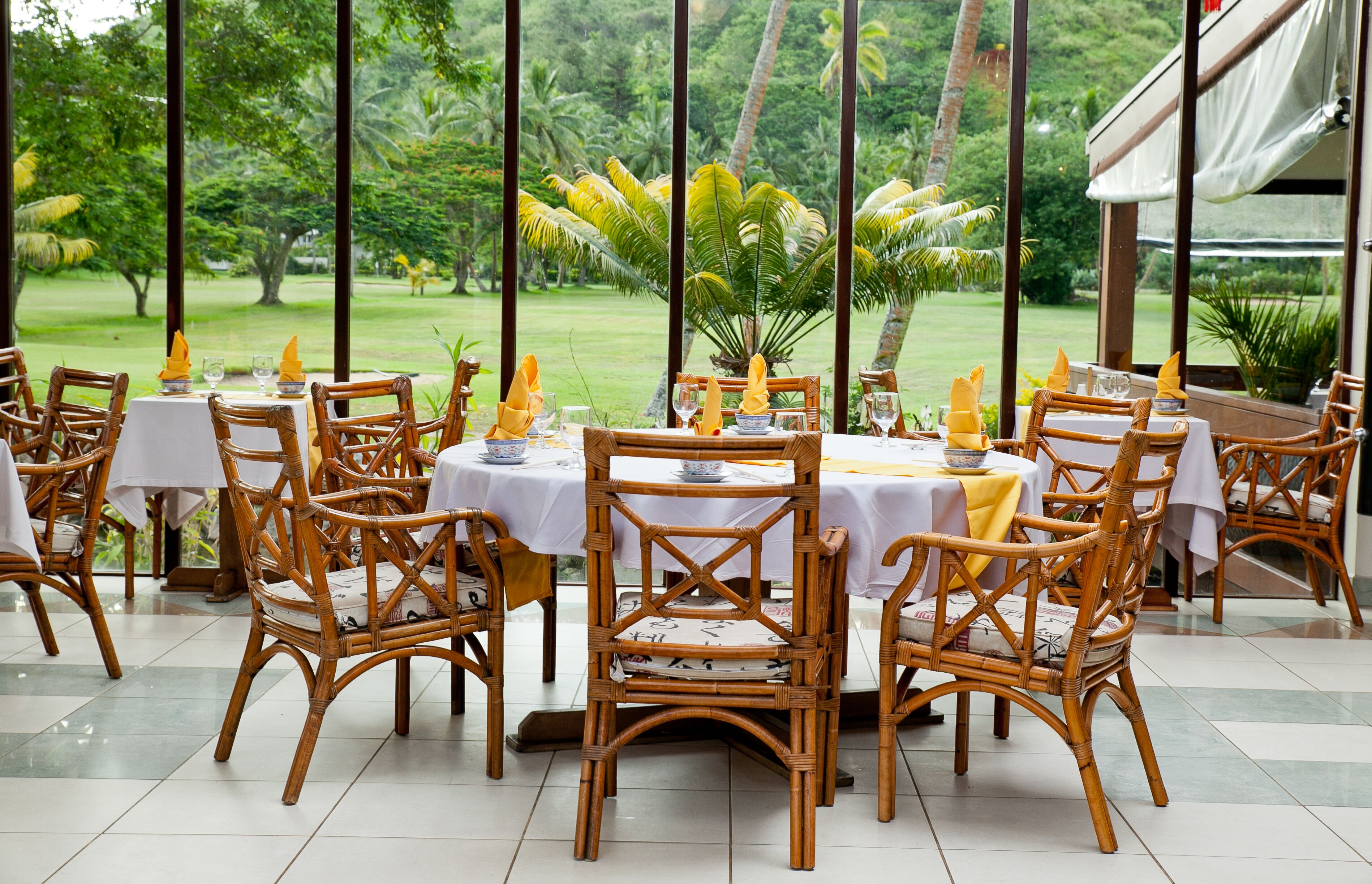Een restaurant met rieten stoelen en witte tafellakens, gelegen tegenover een weelderige tuin.