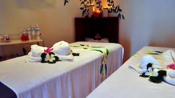 Two beds are dressed with towels and floral arrangements, complemented by a shelf holding toiletries.