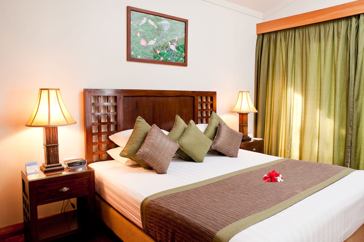 Habitación de hotel impecable con una cama grande, dos lámparas de noche, una mesita de noche con lámpara, un cuadro en la pared y una cortina verde.