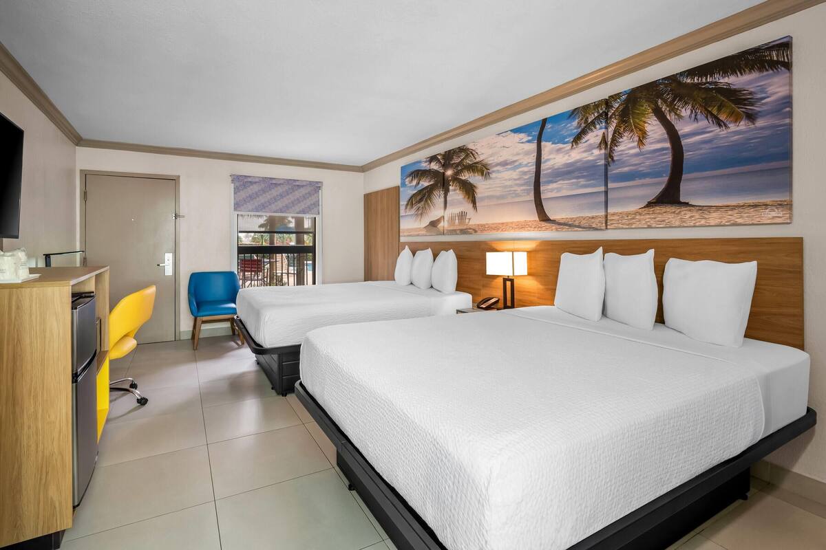 A hotel room with two beds, a desk, a chair, a TV, and a painting of palm trees.