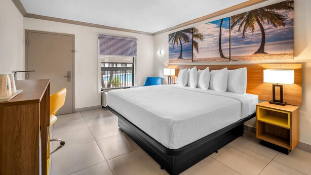 A hotel room with a bed, a desk, a chair, a nightstand, and a painting of a beach scene. - Days Inn by Wyndham Florida City (Florida City)