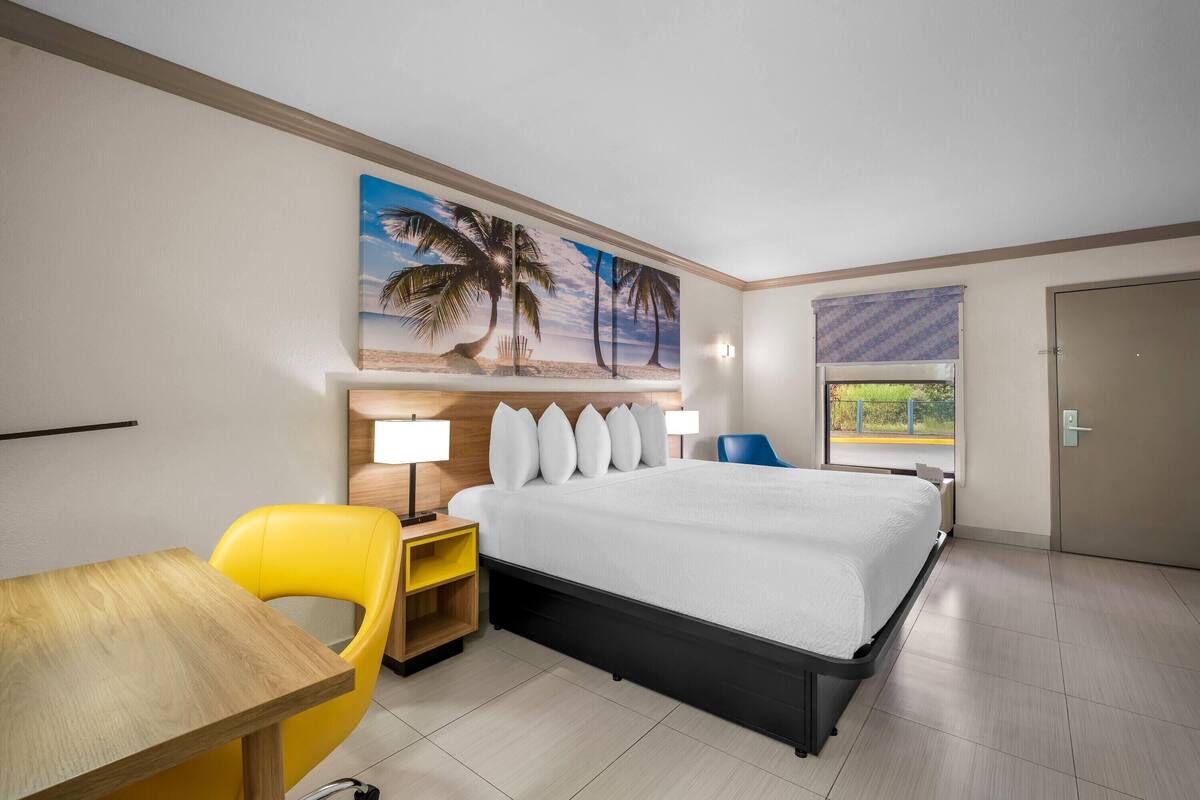 A hotel room with a bed, a desk, a chair, a painting of palm trees, and a window with a view.