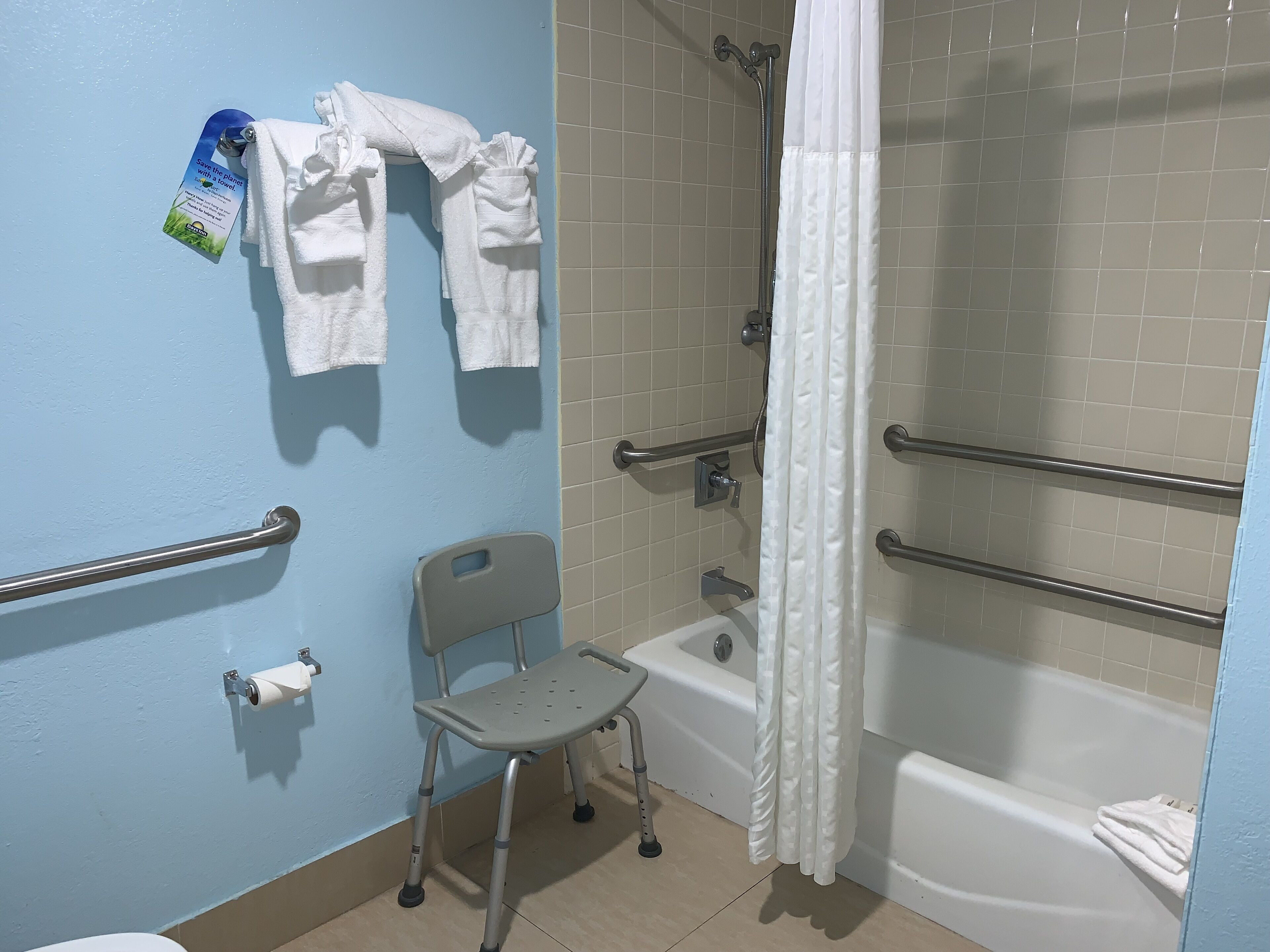 A bathroom with a wheelchair-accessible shower, grab bars, a toilet with a paper towel holder, and a chair.