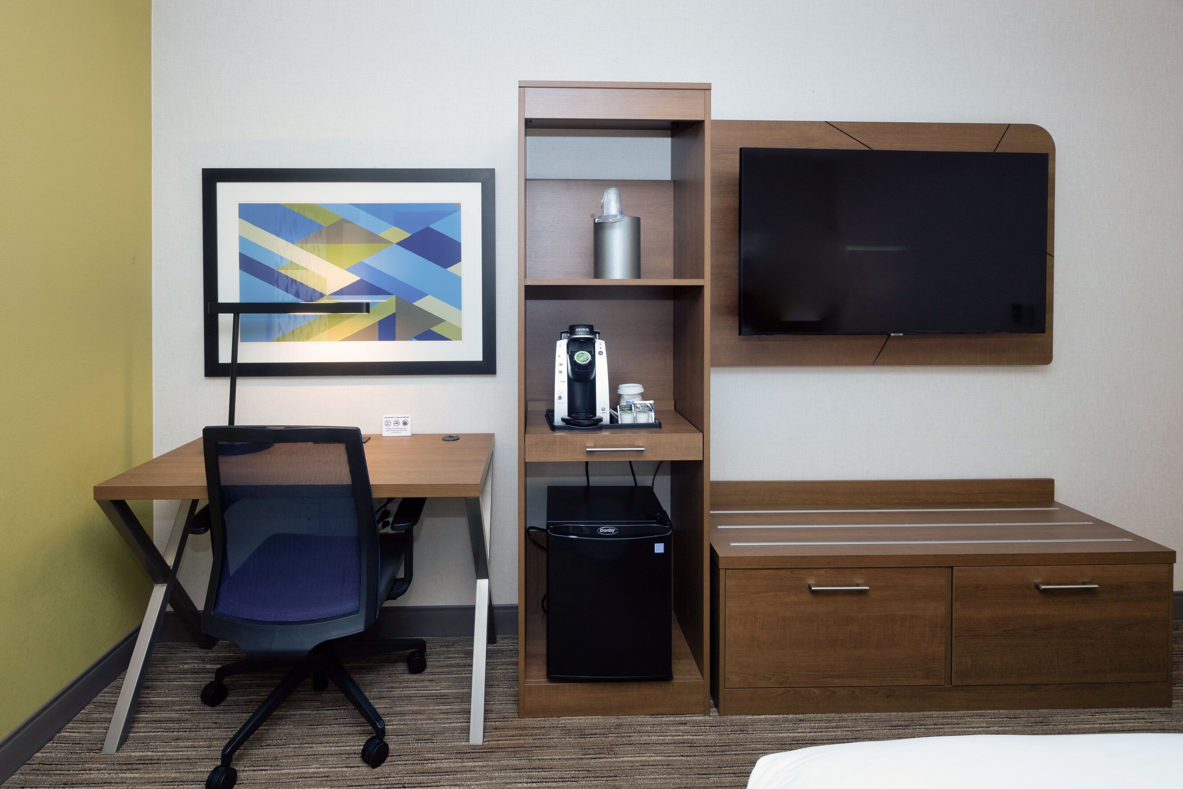 A hotel room with a desk, chair, bed, TV, and a coffee machine.