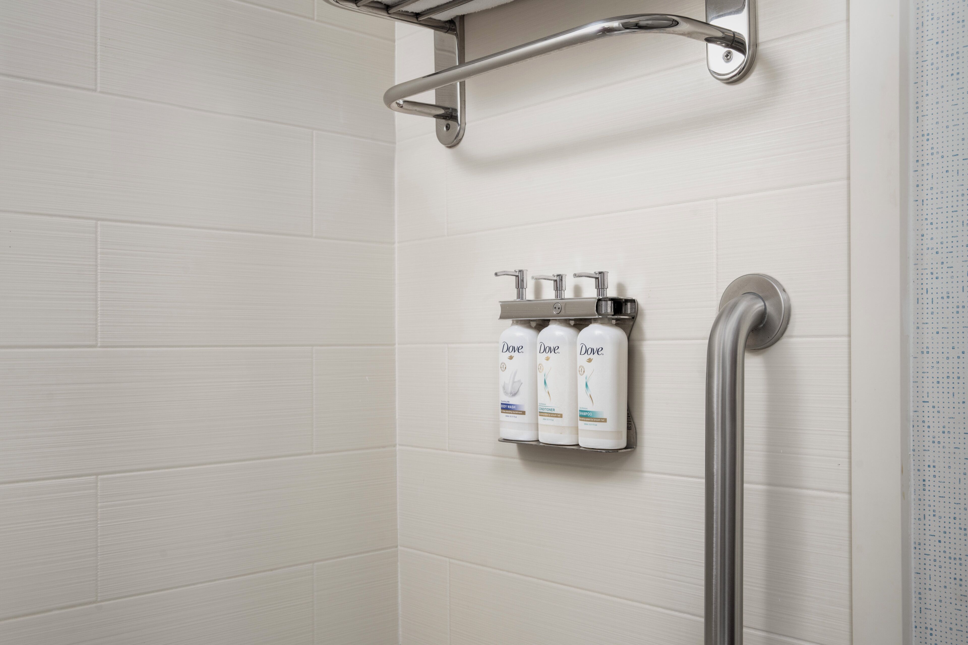 A shower area with white tiled walls, a grab bar, and three Dove shampoo bottles mounted on the wall.