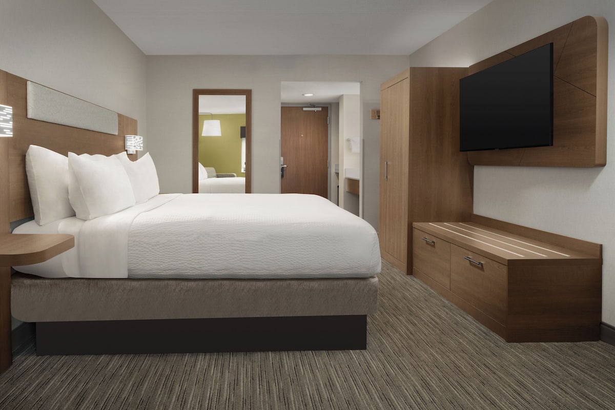 A modern hotel room with a large bed, a bedside table, a TV mounted on the wall, and a wooden wardrobe.