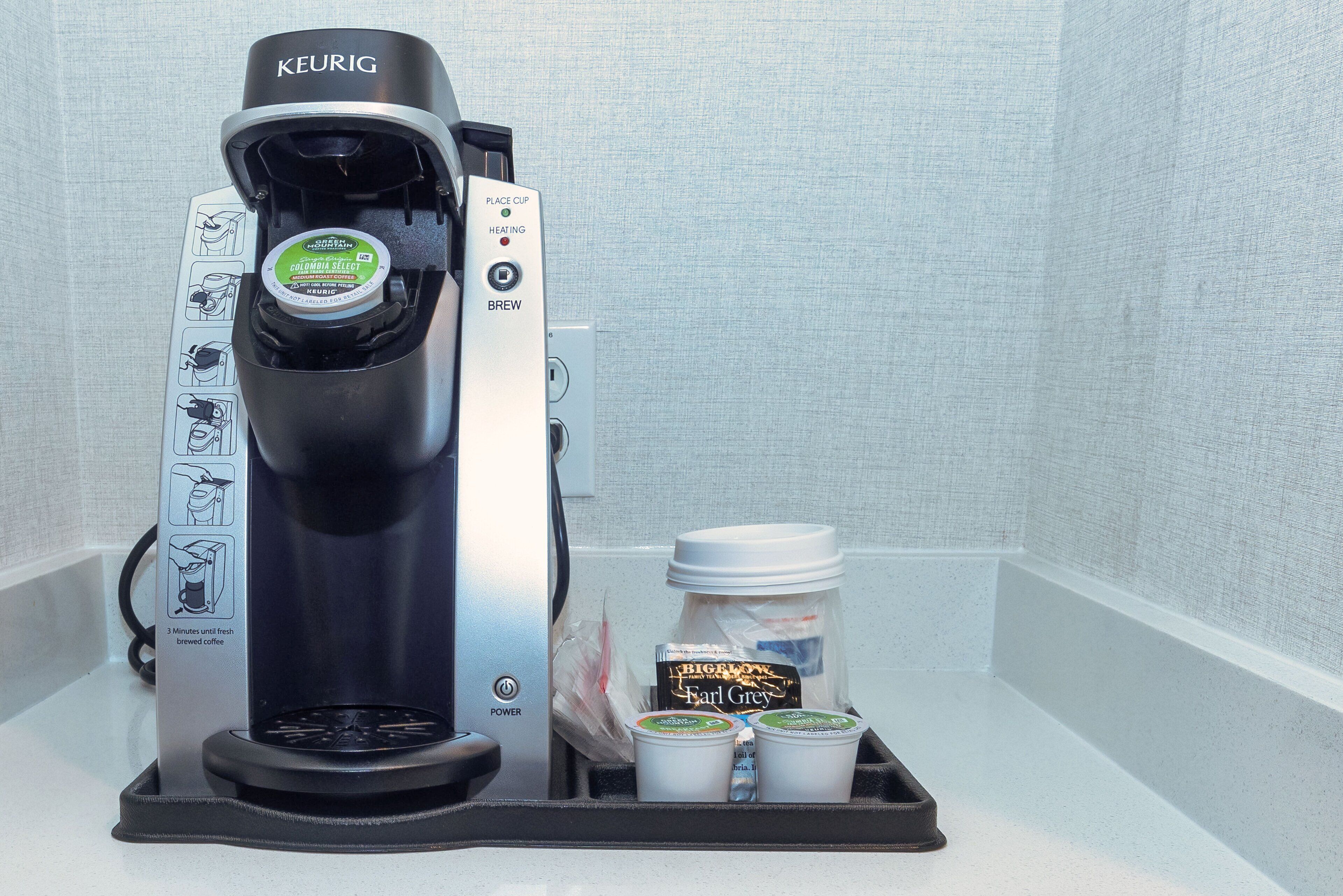 A Keurig coffee maker with a drip tray, a jar of Earl Grey tea, and two disposable cups.
