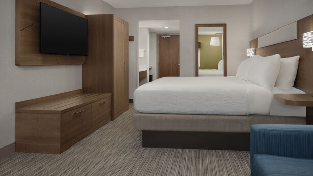 A modern hotel room with a large bed, a TV mounted on the wall, a wooden dresser, and a bedside table with a lamp.