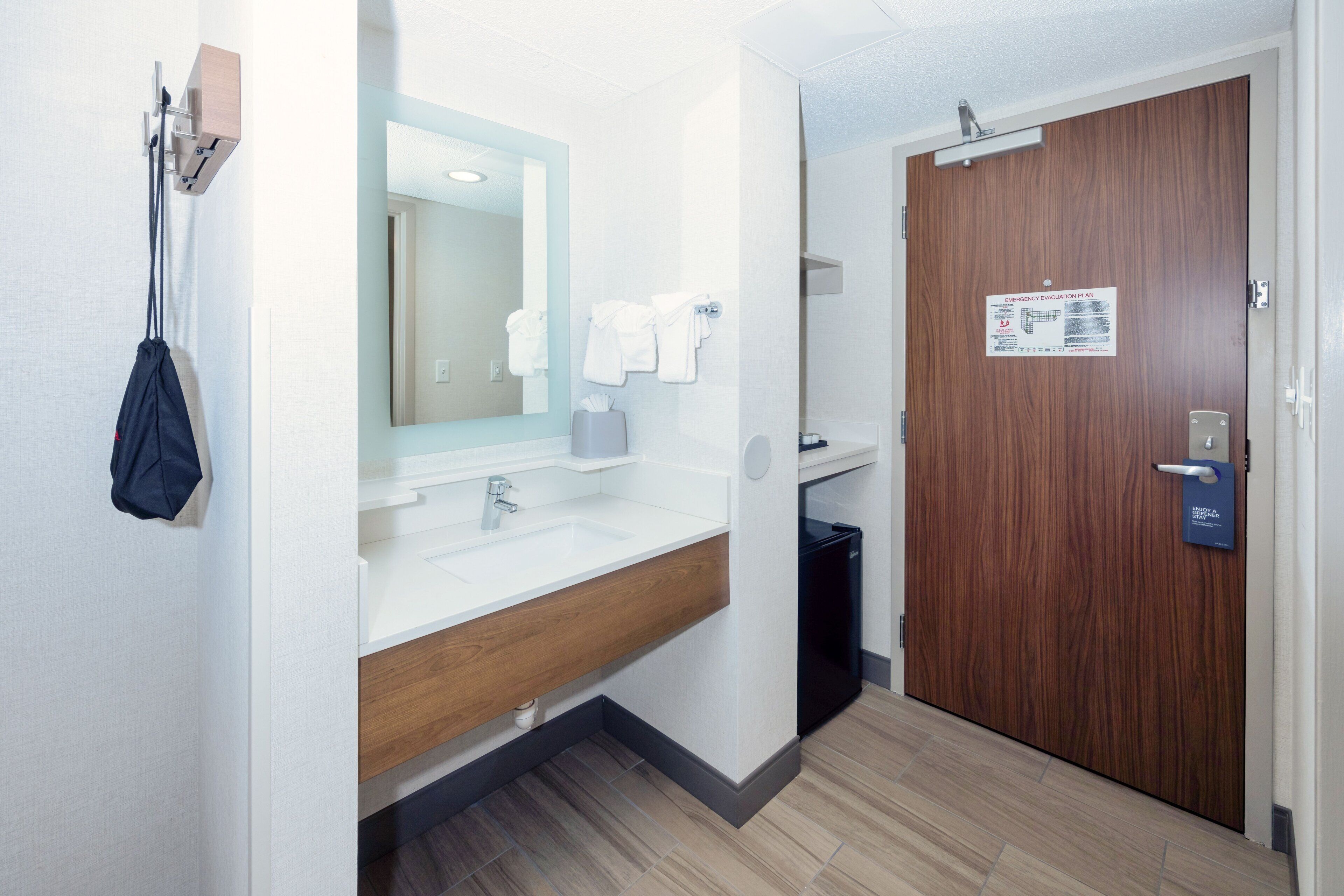 A hotel bathroom with a sink, mirror, towel rack, and a door with an emergency evacuation plan sign.