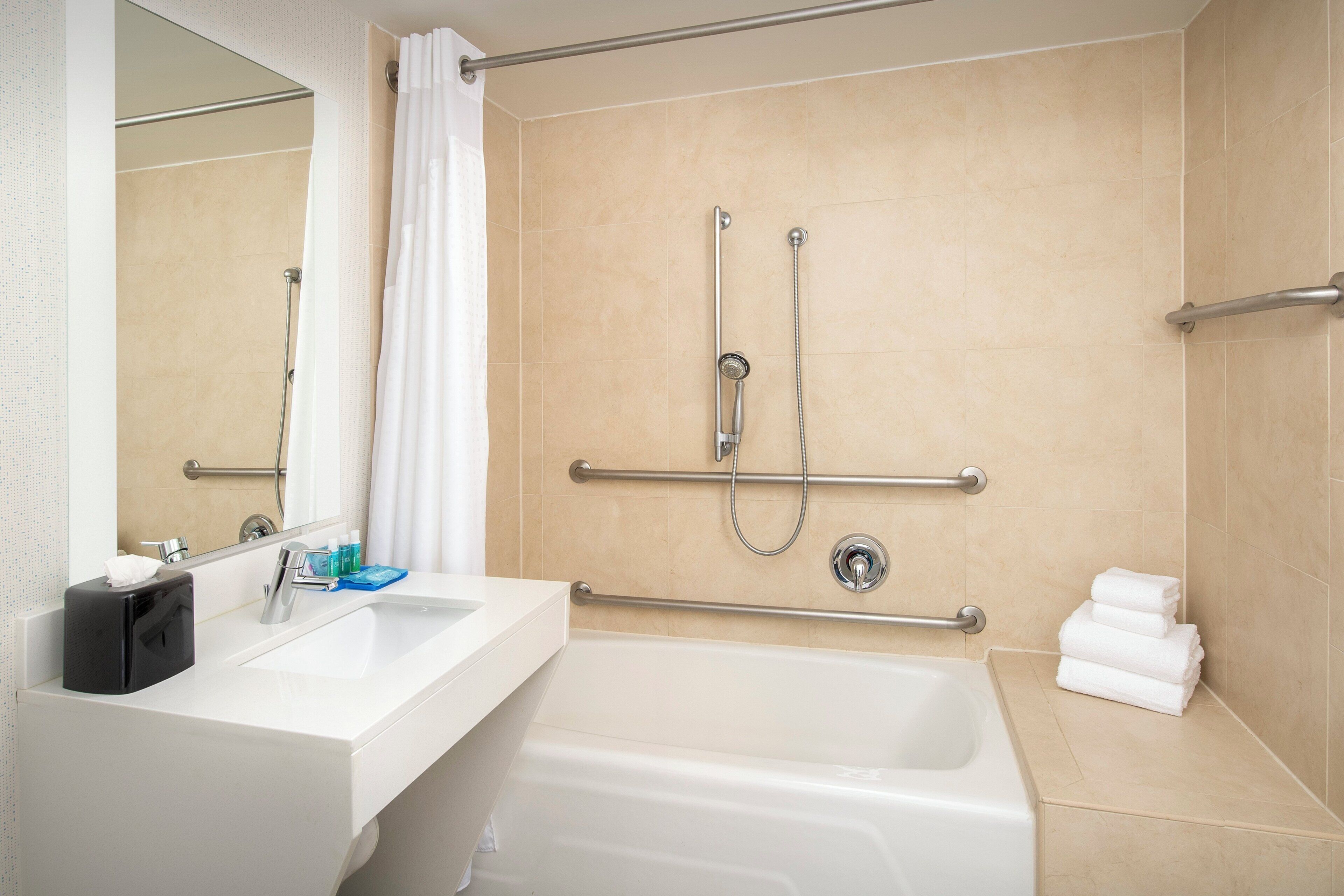 Een moderne badkamer met een bad, een douche met handgrepen, een wastafel met zeepdispenser en een handdoekrek.