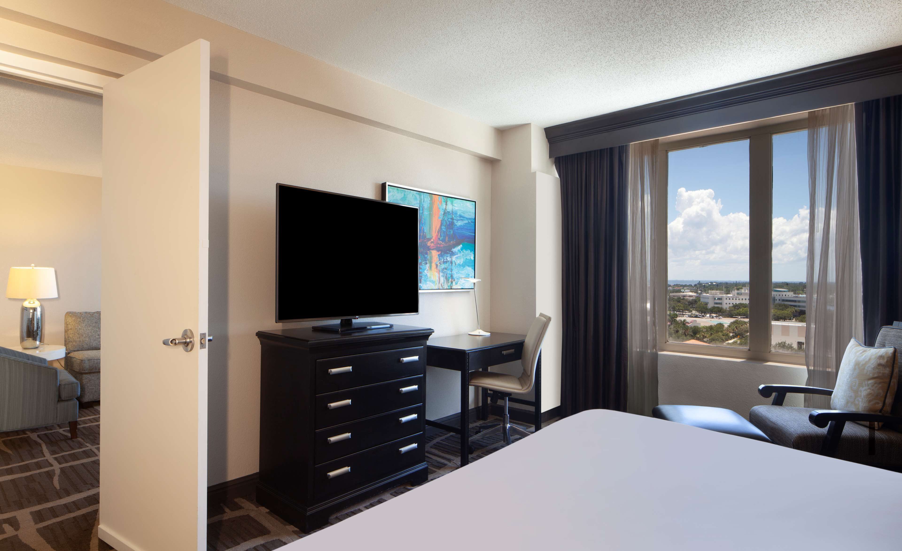 Habitación de hotel con cama, escritorio, silla, televisor y un amplio ventanal con vistas.
