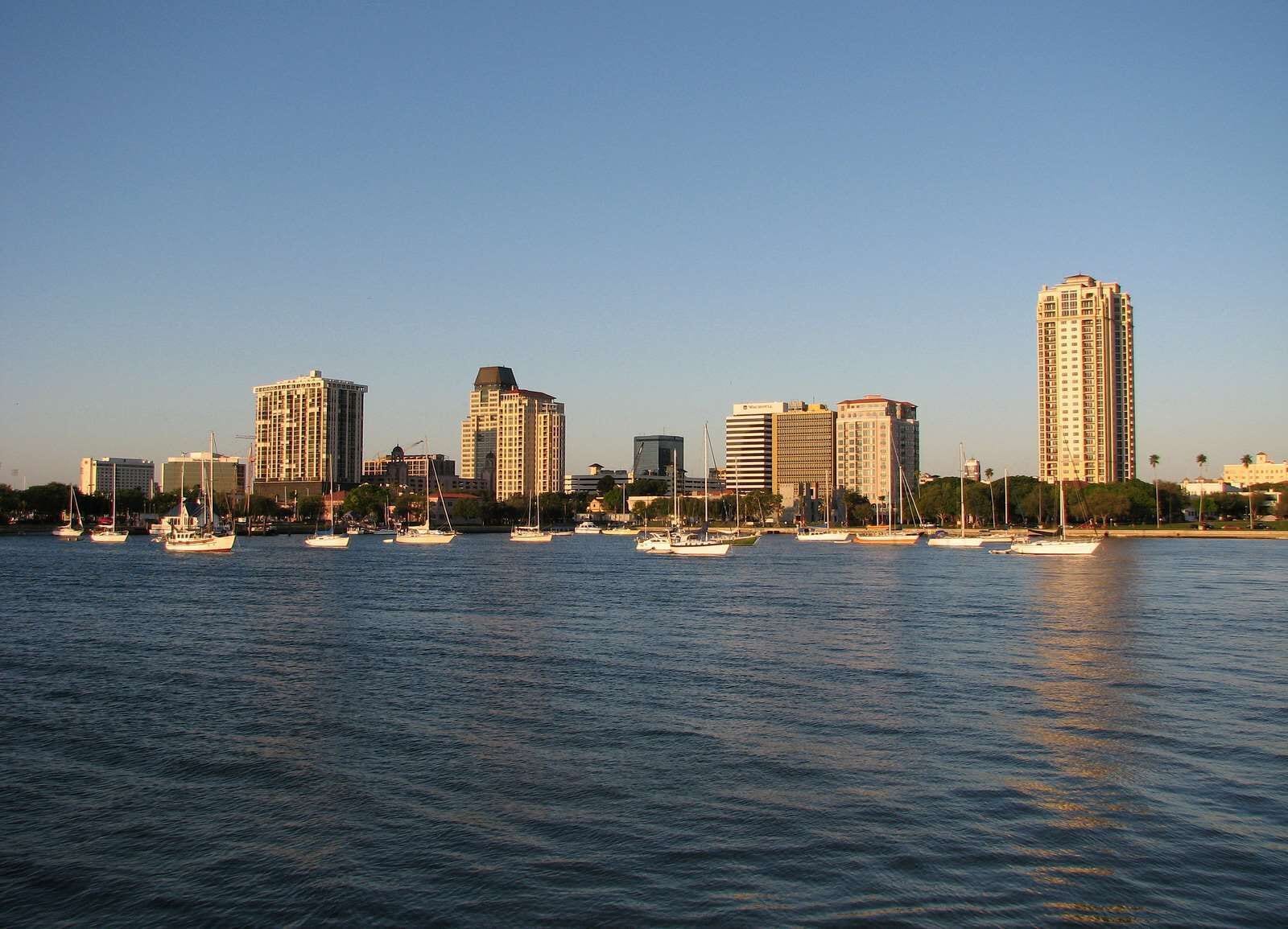 A waterfront cityscape with multiple high-rise buildings and several boats docked along the shore.
