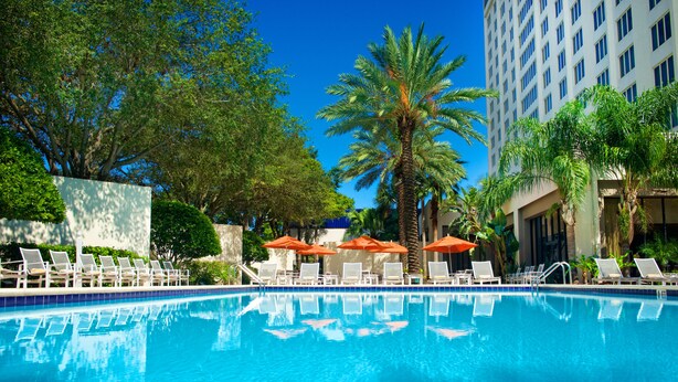 A swimming pool with lounge chairs and umbrellas, surrounded by palm trees and a tall building.