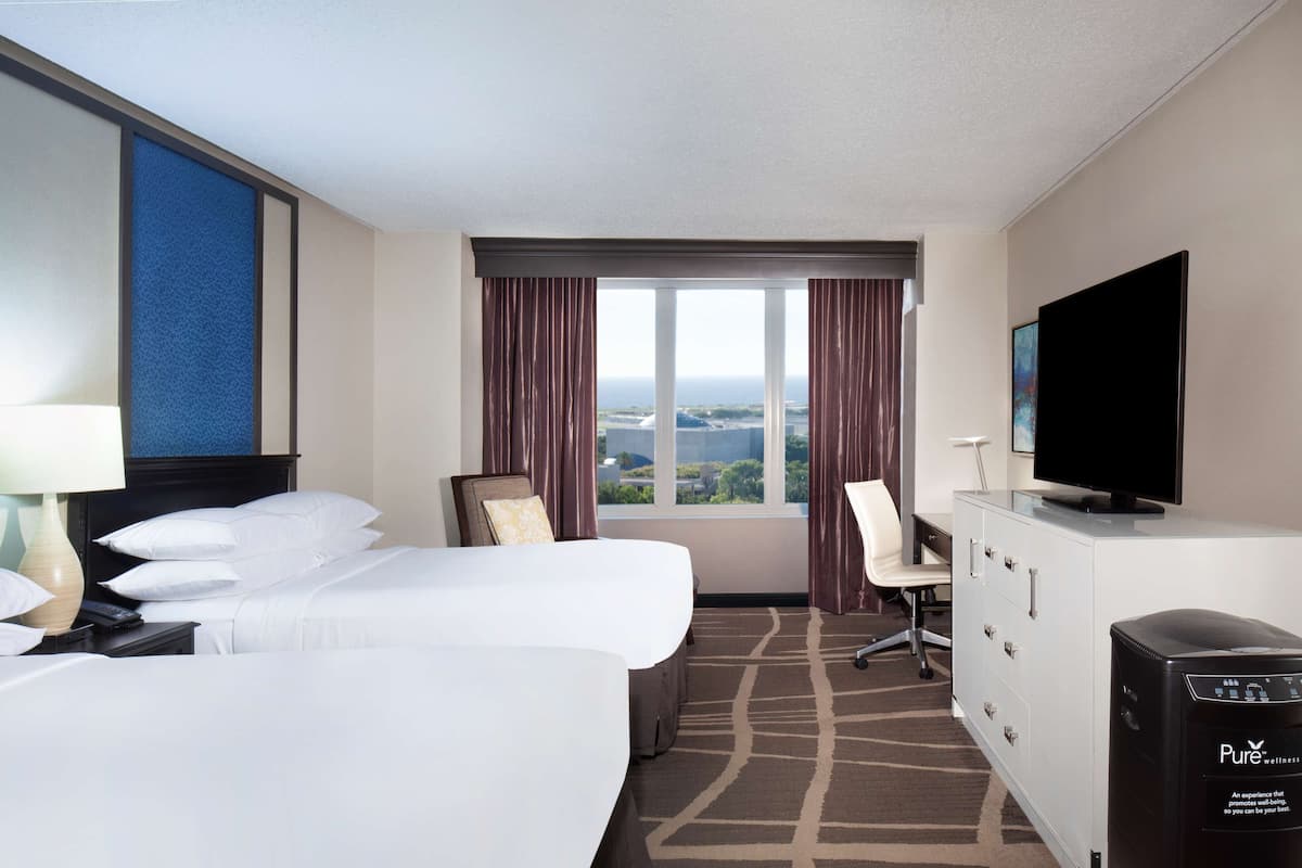 A hotel room with two beds, a TV, a desk, and a view of the city.