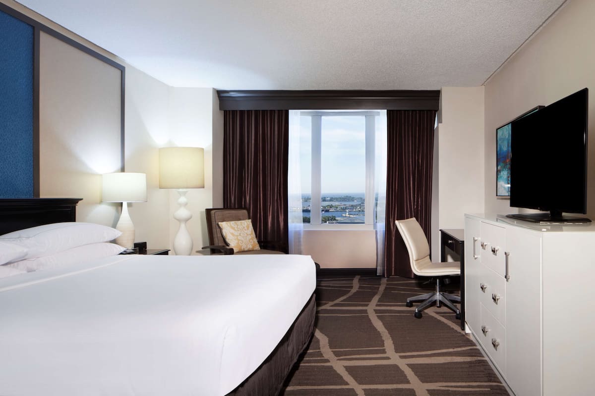 A hotel room with a large bed, a desk with a chair, a television, and a view of the city through the window.
