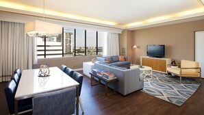 A modern hotel room with a dining area, a sofa, a television, and a view of the cityscape.