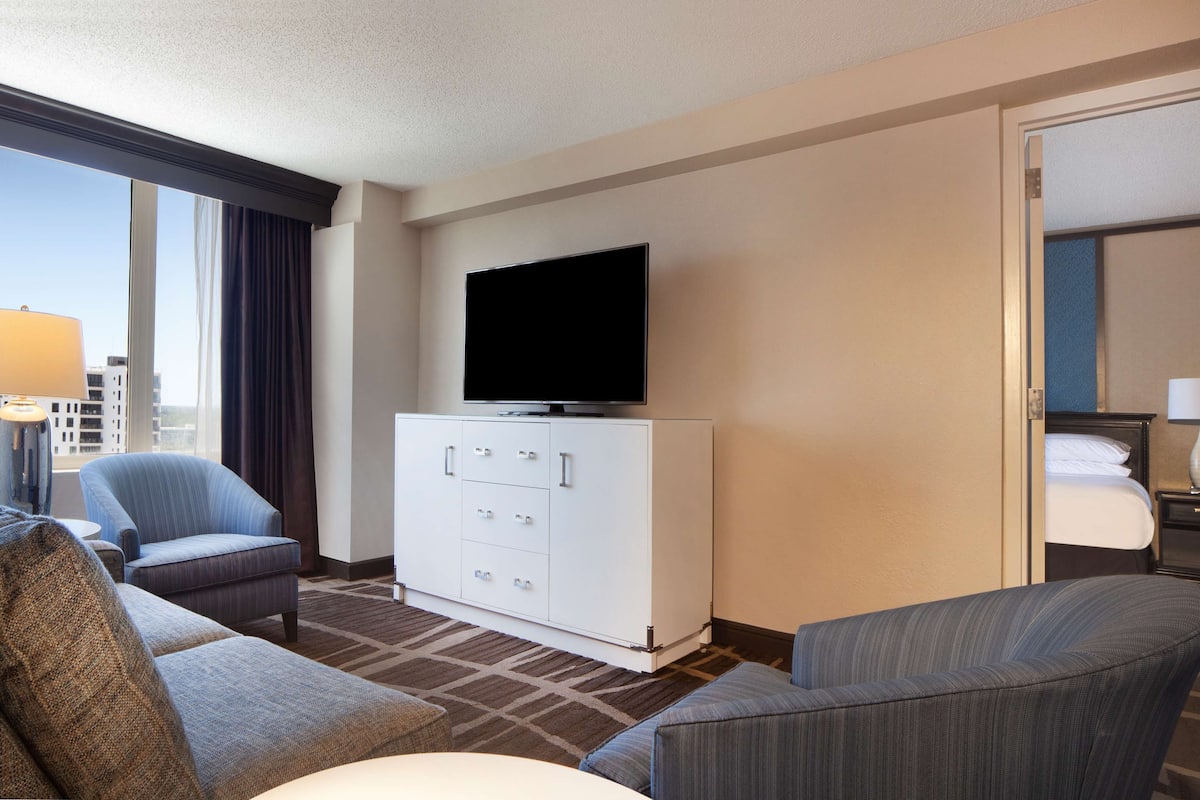 A hotel room with a flat-screen TV on a white cabinet, two armchairs, a sofa, a bed, and a nightstand with a lamp.