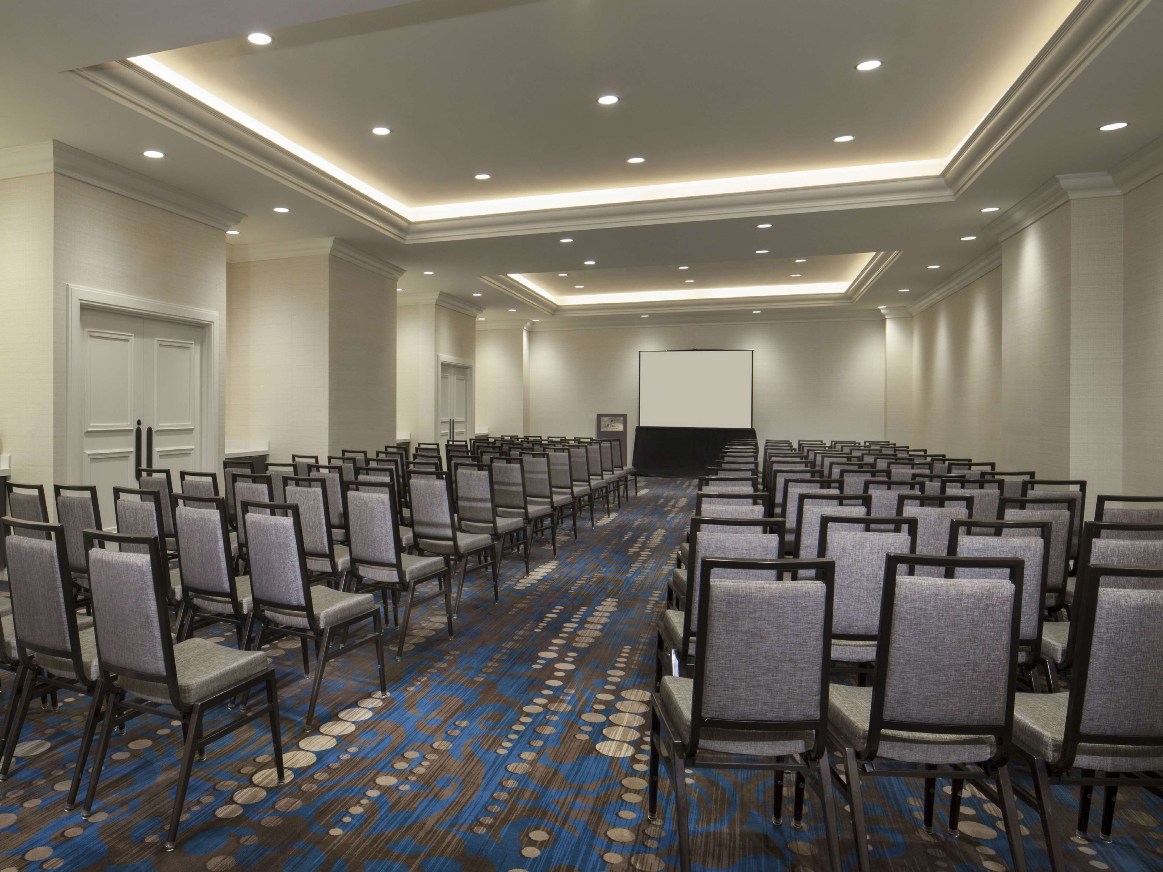 A conference room with rows of chairs, a large screen, and a carpeted floor.