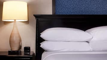 Una cama bien hecha con sábanas y almohadas blancas, una mesita de noche con una lámpara y un pequeño dispositivo electrónico.